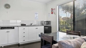 Een kleine keuken met witte kastjes, een magnetron en een spoelbak. Er staat een eettafel met stoelen en een raam met jaloezieën.