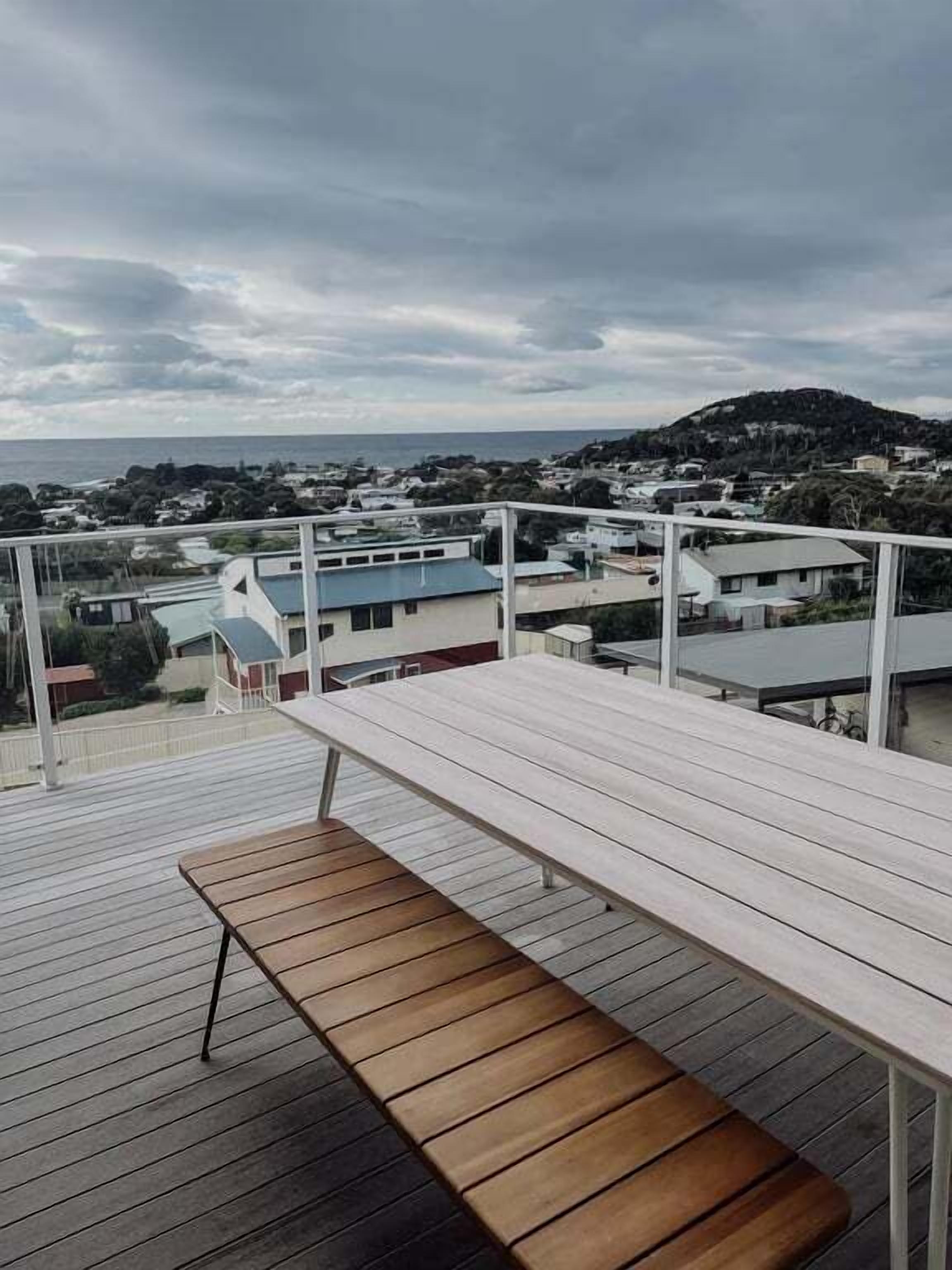 A balcony with a wooden bench, overlooking a coastal town with buildings and a hill in the distance.