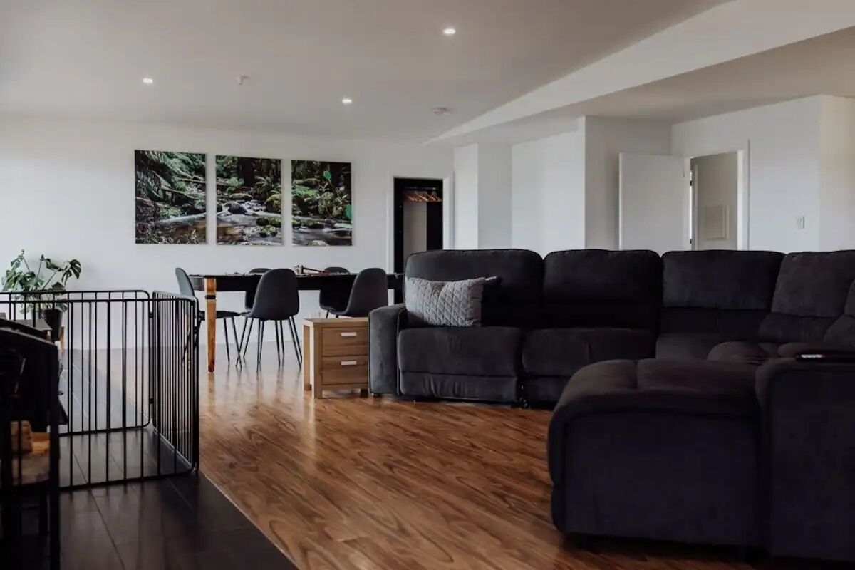 Een moderne woonkamer met een zwarte hoekbank, een eethoek met een tafel en stoelen, en uitzicht op een bos door grote ramen.