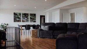 A modern living room with a black sectional sofa, a dining area with a table and chairs, and a view of a forest through large windows. - Bayview Lodge - Views Views Views! (Break O'Day)