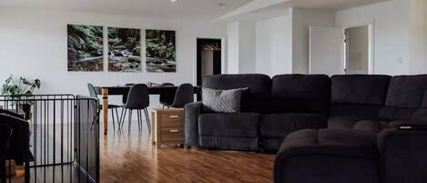Een moderne woonkamer met een zwarte hoekbank, een eethoek met een tafel en stoelen, en uitzicht op een bos door grote ramen.