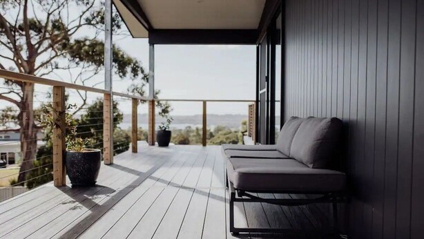 A balcony with a wooden floor, a grey sofa, and a potted plant. - Bayview Lodge - Views Views Views! (Break O'Day)
