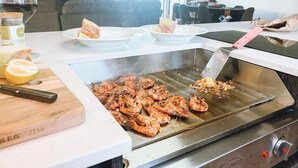 A kitchen with a grill cooking shrimp, a cutting board with lemons, and a wine glass. - Bayview Lodge - Views Views Views! (Break O'Day)