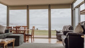 A room with a large sliding glass door leading to a view of the sea, a sofa, a dining table with chairs, and a small wooden table. - OceanFront - Absolute Waterfront (Glamorgan-Spring Bay)