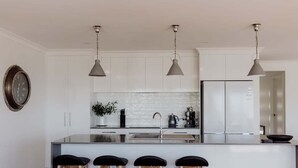A modern kitchen with white cabinetry, a central island with bar stools, pendant lighting, and a stainless steel refrigerator. - Anchored@Bicheno - Ohmyview! (Glamorgan-Spring Bay)