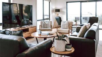 A modern living room with a large flat-screen TV, comfortable seating, and large windows offering a view of the outdoors.