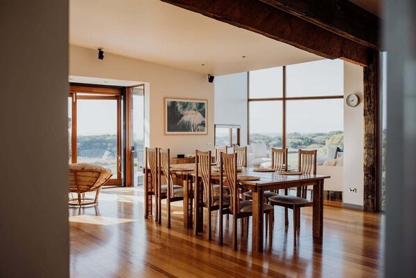 A dining area with a wooden table and chairs, large windows with a view, and a painting on the wall.