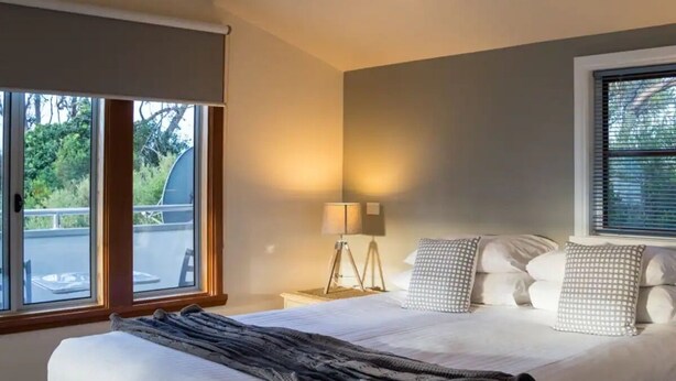 Un dormitorio con una cama grande, una mesita de noche con lámpara, una ventana con persianas y vistas al exterior.