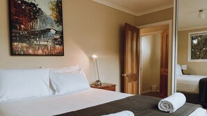 A hotel room with a large bed, a painting on the wall, a nightstand with a lamp, and a door leading to another room. - Barefoot Bungalow | Cozy Bicheno Hideaway | (Glamorgan-Spring Bay)