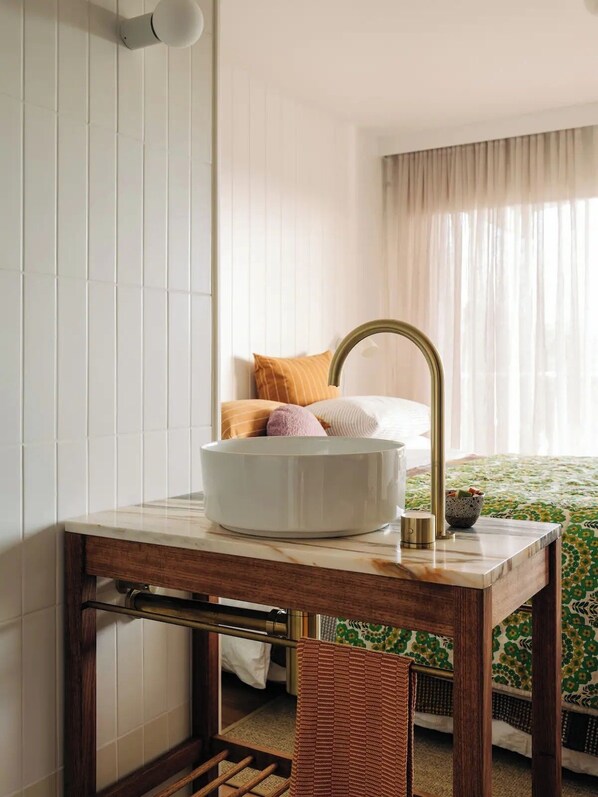 Une salle de bain avec un lavabo blanc, un robinet doré et un miroir. On y trouve un lit recouvert d’une couvre-lit à motifs floraux verts et une chaise marron.