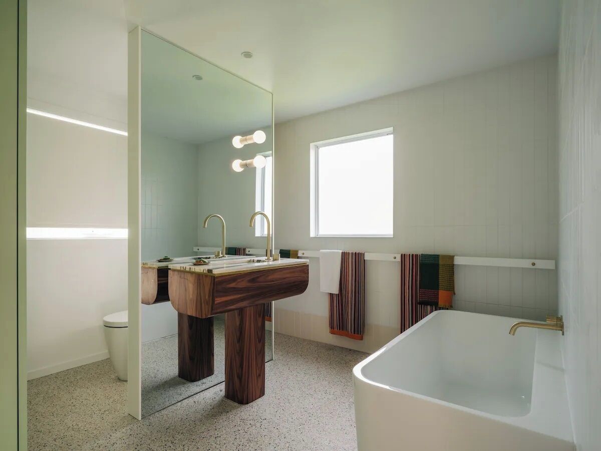Ein modernes Badezimmer mit Waschtisch aus Holz, einem großen Spiegel, einer Badewanne und einem Fenster für natürliches Licht.