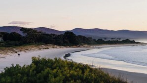 A beach with a person walking along the shore, surrounded by hills and sparse vegetation. - Frankie Beach House Bicheno (Glamorgan-Spring Bay)