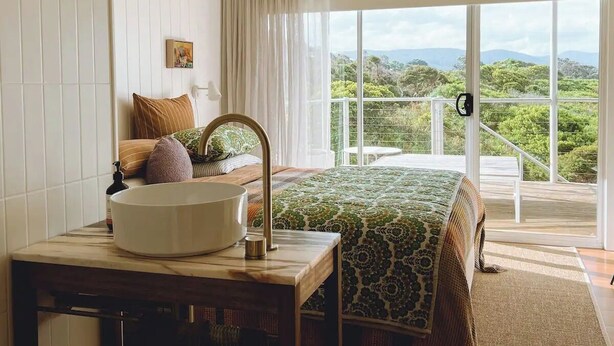 A bedroom with a large window, a bed, a bedside table with a sink, and a view of greenery outside. - Frankie Beach House Bicheno (Glamorgan-Spring Bay)