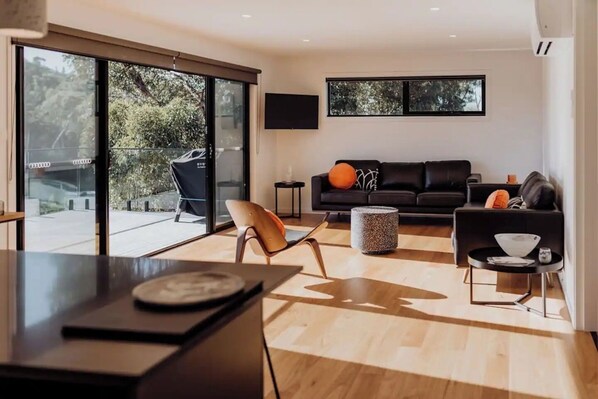 Un salon moderne avec un canapé noir, une table à manger en bois et une porte-fenêtre en verre qui donne sur une terrasse.