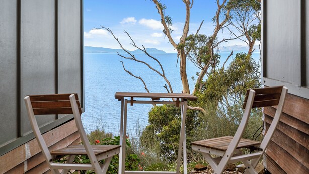 A balcony with two wooden chairs and a table, overlooking a body of water and distant mountains.