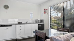 A modern kitchen with white cabinets, a microwave, and a dishwasher. There is a dining table with chairs and a window with blinds.
