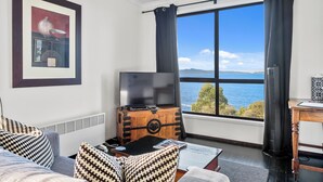 A room with a large window, a television on a wooden chest, a sofa with patterned cushions, and a framed picture on the wall.