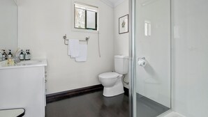 A bathroom with a white toilet, a glass shower enclosure, a white sink with soap dispensers, and a window above the toilet.