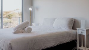 Una cama bien hecha con sábanas blancas, una lámpara de noche y vistas por una ventana grande.