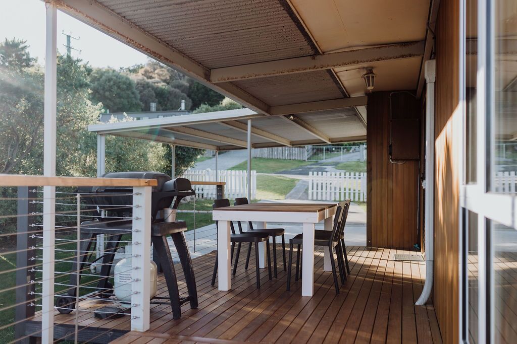 An outdoor patio with a wooden table and chairs, a grill, and a view of a white picket fence and hills in the background.