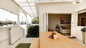 Un área de comedor exterior moderna con una mesa de madera, un tapete verde y vistas a la calle y la vegetación.