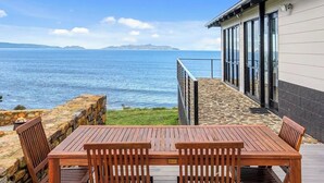 Una terraza de madera con mesa y sillas, con vistas a un cuerpo de agua y una masa terrestre distante.