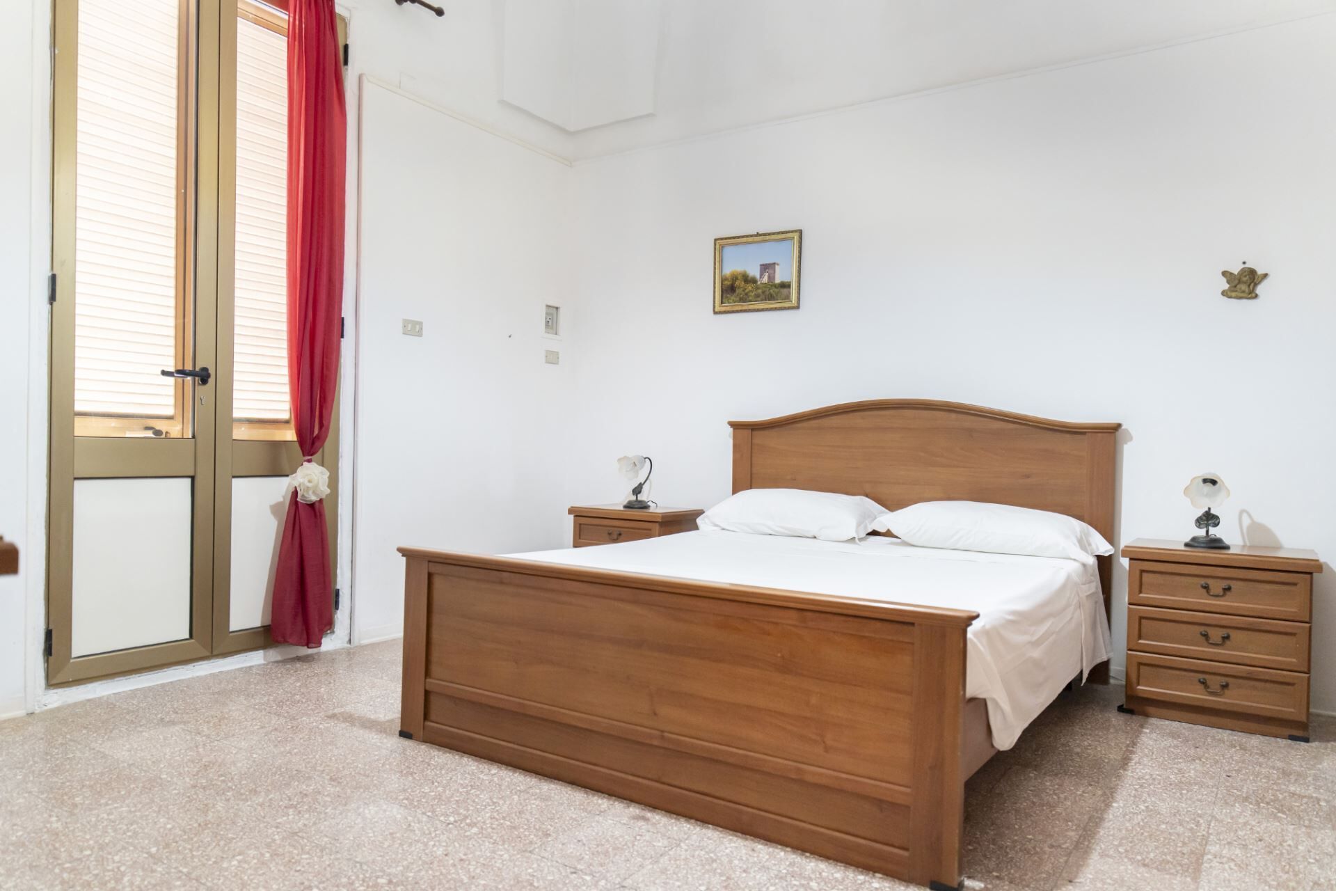 A bedroom with a wooden bed, two bedside tables, a nightstand, a red curtain, a window, and a small picture on the wall.