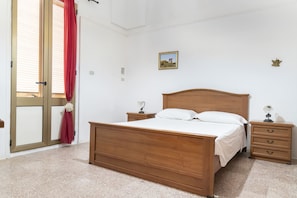 Un dormitorio con una cama de madera, dos mesitas de noche, una mesita auxiliar, una cortina roja, una ventana y un cuadro pequeño en la pared.