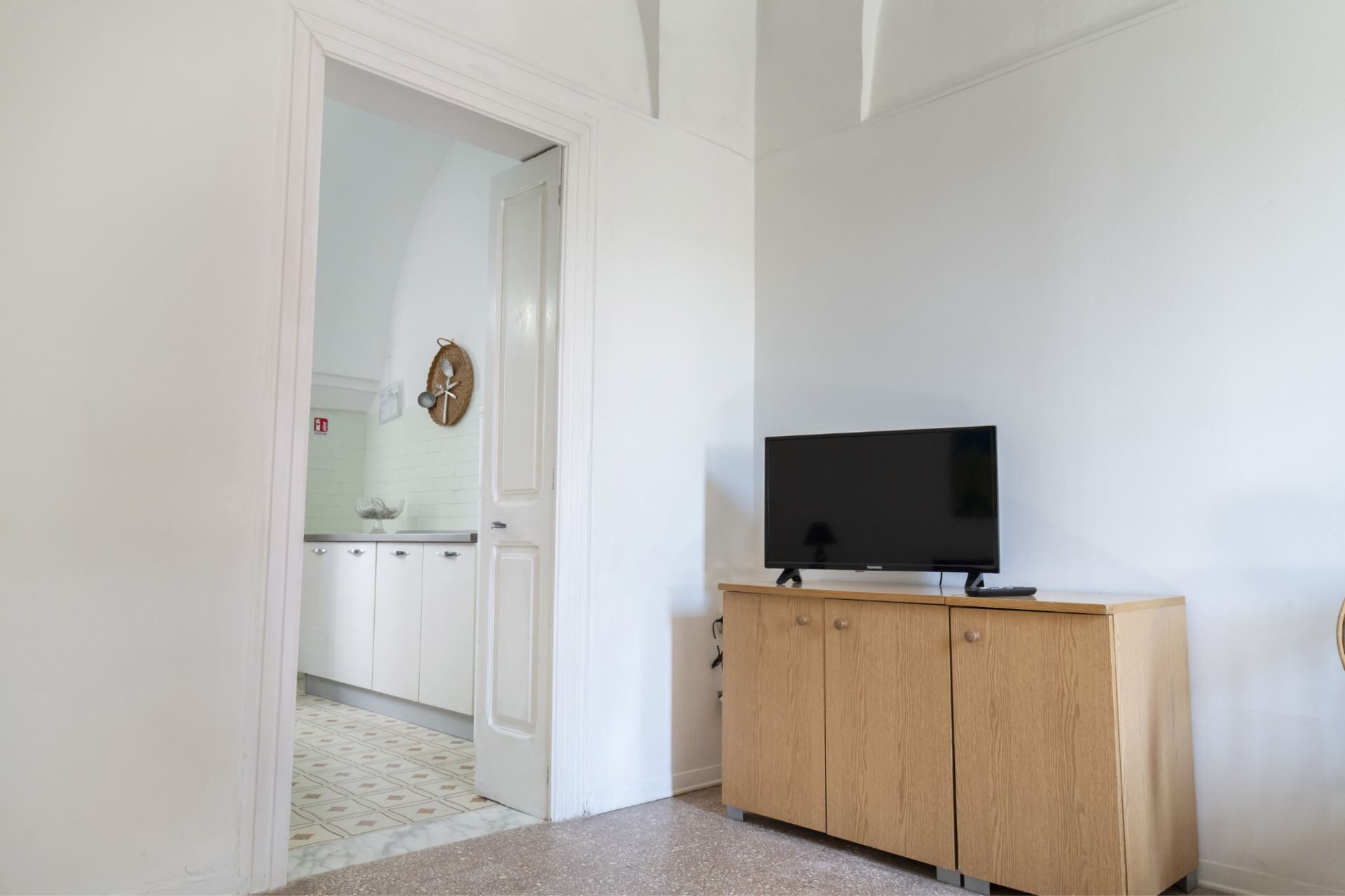 A room with a television on a wooden cabinet, a white door leading to a kitchen, and a tiled floor.