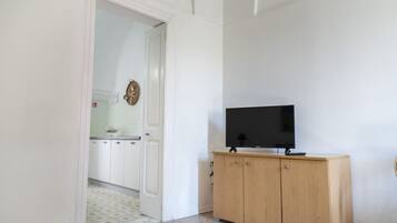 A room with a television on a wooden cabinet, a white door leading to a kitchen, and a tiled floor.