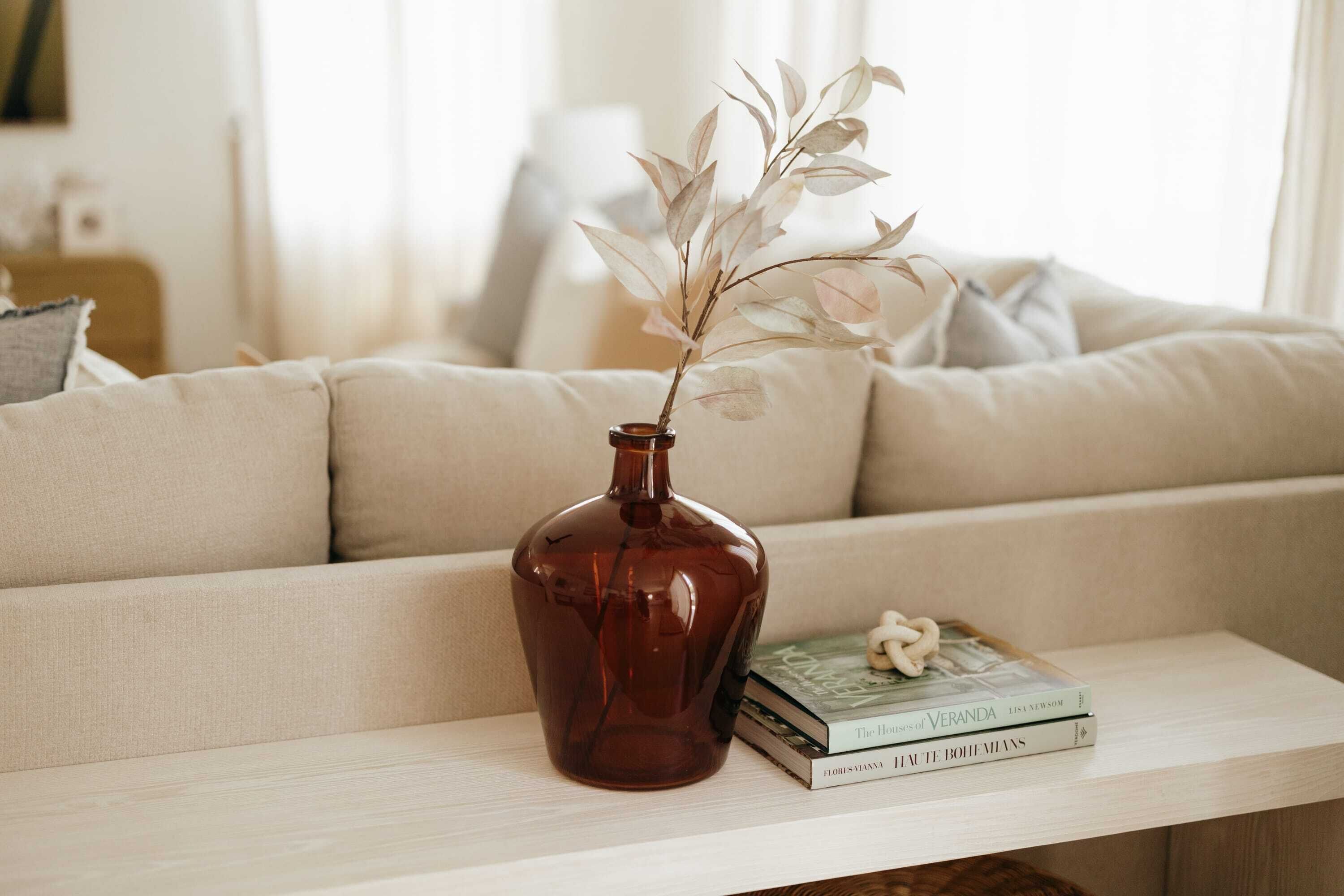 Eine braune Vase mit getrockneten Blättern auf einem weißen Tisch neben einem beigefarbenen Sofa.