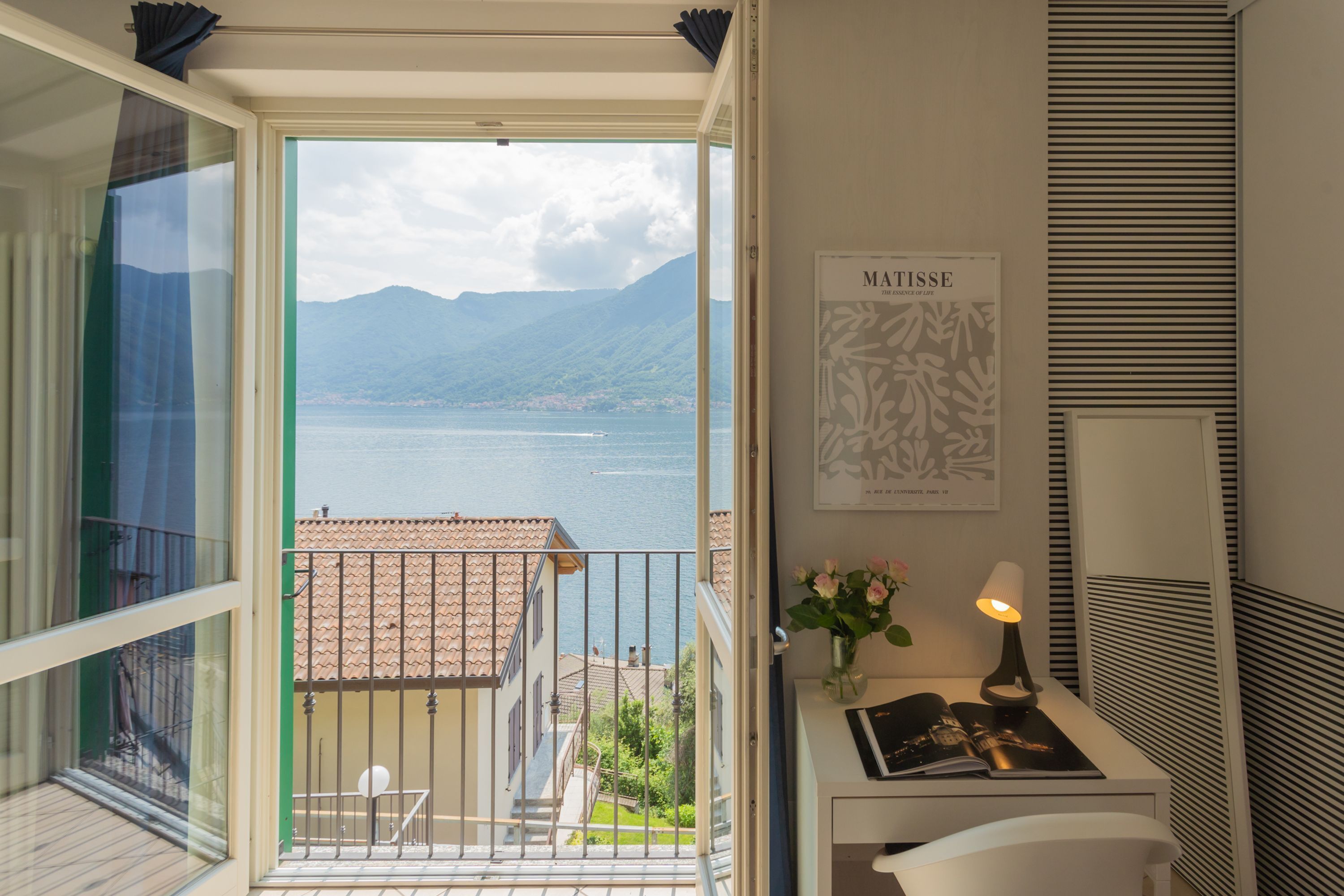 A balcony with a view of a lake and mountains, a framed artwork, a lamp, and a small table with a book and flowers.