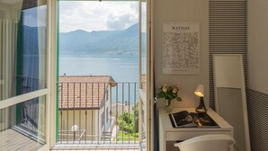 Un balcon donnant sur un lac et des montagnes, une œuvre d’art encadrée, une lampe et une petite table avec un livre et des fleurs.