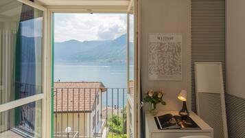 Un balcón con vista a un lago y montañas, una obra de arte enmarcada, una lámpara y una pequeña mesa con un libro y flores.