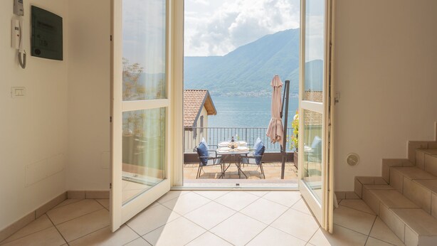 Une porte ouverte donnant sur un balcon avec une table et des chaises, une vue sur la mer et une montagne en arrière-plan.