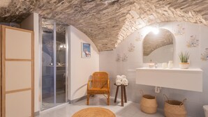 Une salle de bain avec un plafond en pierre, une porte de douche en verre, un fauteuil en osier, un lavabo avec un miroir et une plante en pot.
