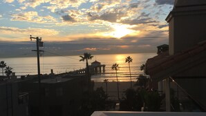 Een zonsondergang uitzicht vanaf een balkon, met uitzicht op een strand met palmbomen en een pier.