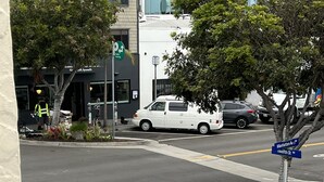 Een straatoversteekplaats met een blauwe gevelpad, een witte bestelwagen en een blauw straatnaambordje waarop 'Manhattan Ave' en 'Welfield St' staan.