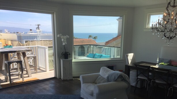 Een ruime woonkamer met een balkon met uitzicht op zee, een eettafel met stoelen en een kroonluchter.