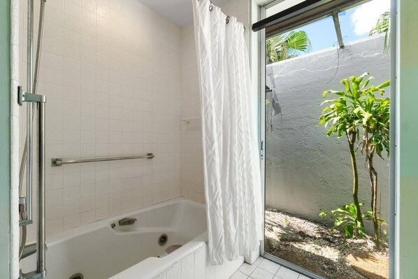 Un baño con bañera, ducha y puerta de vidrio que da a un balcón con vistas a una zona rocosa y un árbol pequeño.