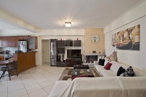 A living room with a sofa, a coffee table, and a fireplace. There is a kitchen area with cabinets and a refrigerator. - Paradeisos in Chalandri (Marousi)