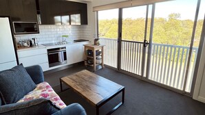 En moderne opholdsstue med et køkken, spiseplads og en balkon med udsigt til træer.