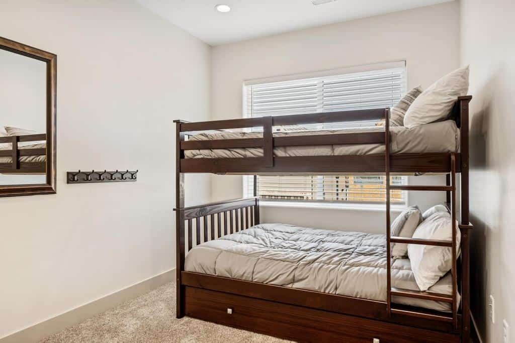 A bunk bed with a wooden frame, a window with blinds, and a mirror on the wall.