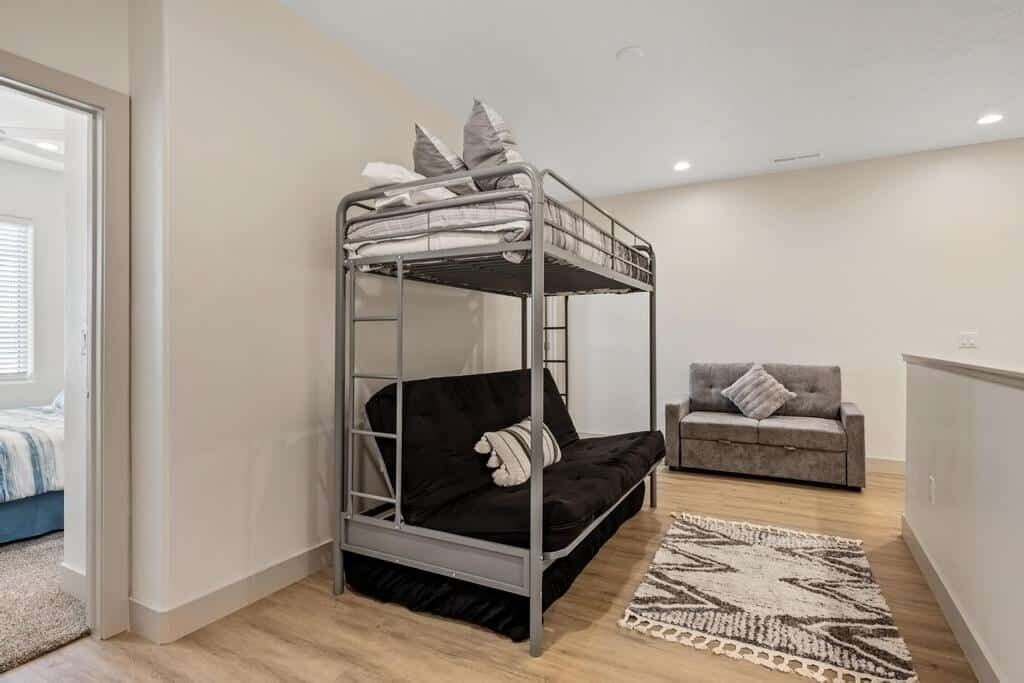 Een stapelbed met kussens, een grijze bank en een kamer met houten vloeren en witte muren, met een patroonkleed.