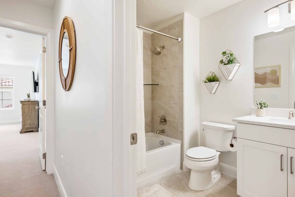 Een moderne badkamer met bad, toilet en douche. Er staan planten aan de muur en er hangt een spiegel boven de wastafel.