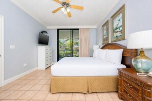 A hotel room with a bed, a ceiling fan, a TV, a dresser, and a view of the outdoors.