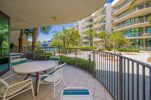 A balcony with a round table and chairs, a pool area with lounge chairs, and a multi-story building with balconies.