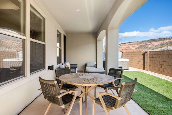 A covered patio with a round wooden table and wicker chairs, a white sofa, and a small table with decorative items.