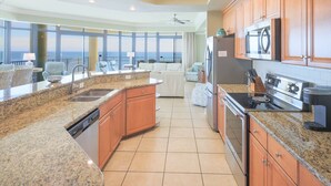 A spacious kitchen with granite countertops, wooden cabinets, and a large island. It features a built-in dishwasher, a stove, and a refrigerator. - Phoenix West 609 4 Bedroom Condo (Orange Beach)