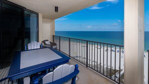 Ein Balkon mit Tisch und Stühlen, der Blick auf einen Strand mit Sonnenschirmen und Menschen freigibt.